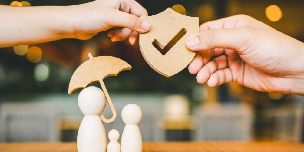 Hands holding a wooden shield with a checkmark and an umbrella over a family of wooden figures, visually demonstrating the importance of protection and why you need renters insurance to safeguard your belongings.