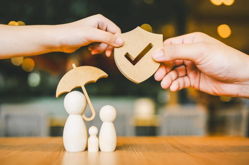 Hands holding a wooden shield with a checkmark and an umbrella over a family of wooden figures, visually demonstrating the importance of protection and why you need renters insurance to safeguard your belongings.
