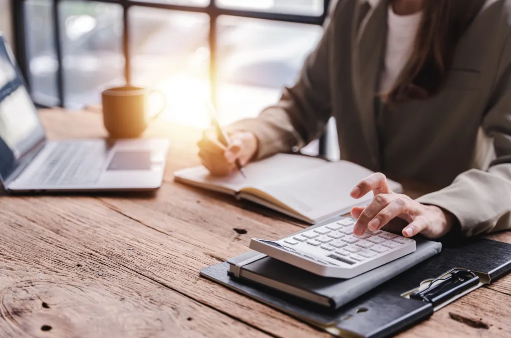Person sitting at a wooden desk using a calculator and writing in a notebook, with a laptop and coffee cup nearby, calculating a renters insurance deductible.