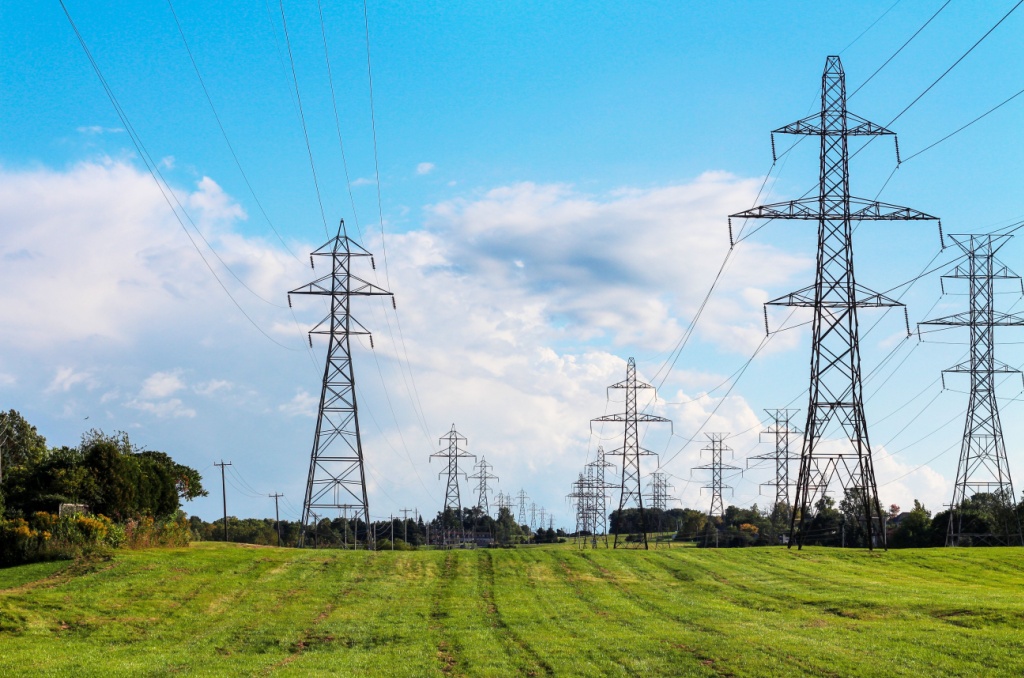 A row of tall electricity transmission towers stretches across a green field under a bright blue sky, illustrating a step in the process of how electricity works.