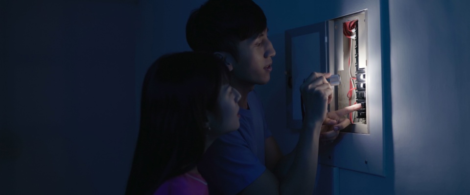Couple using a flashlight to inspect a circuit breaker panel, troubleshooting electrical issues and showing what to do when the power goes out.