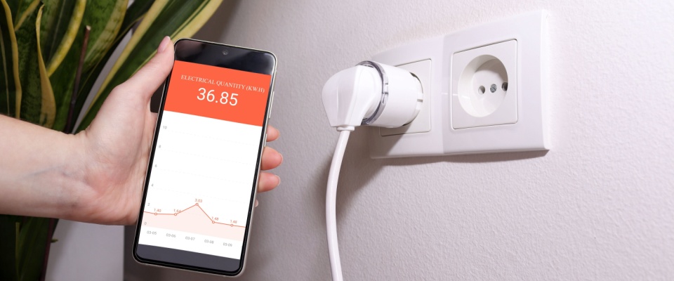 Hand holding a phone showing electricity consumption data next to a smart plug, demonstrating tools commonly used during a home energy audit.
