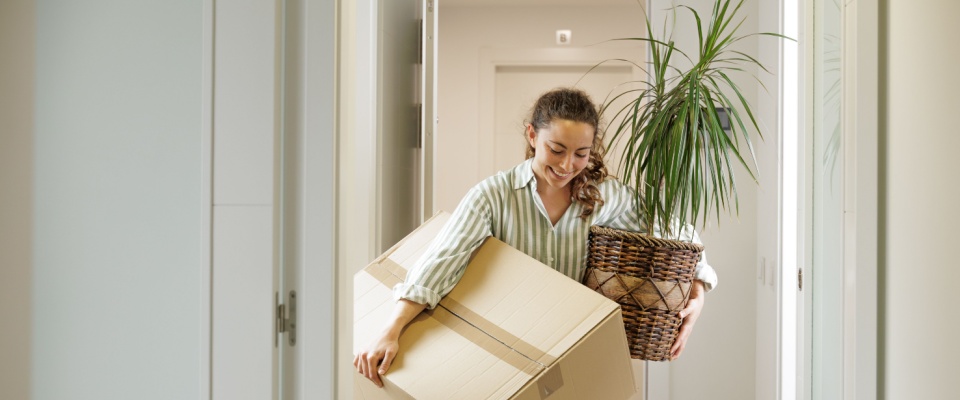 Woman carrying moving boxes and a houseplant into a new home, illustrating key differences between homeowners versus renters insurance when relocating.