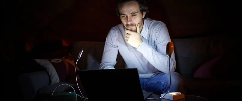 Man working on a laptop by dim light during a rolling blackout, illustrating how temporary outages can disrupt daily routines.