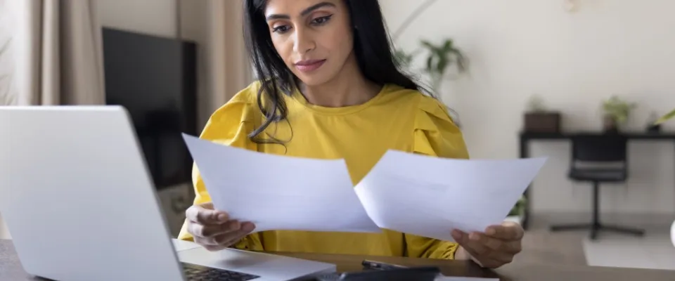 Woman in a yellow blouse reviewing paperwork and comparing documents at her laptop while researching month-to-month rentals from home.