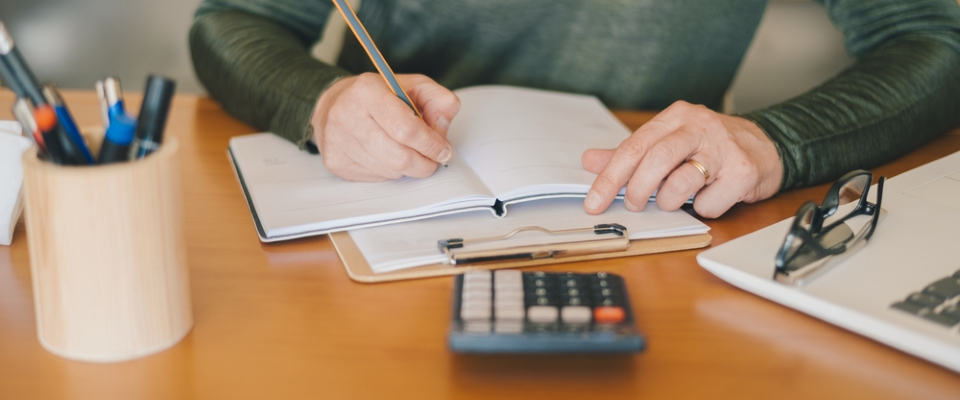 Person writing in a notebook with a calculator, pens, and glasses on a desk while figuring out how to budget for utilities each month.